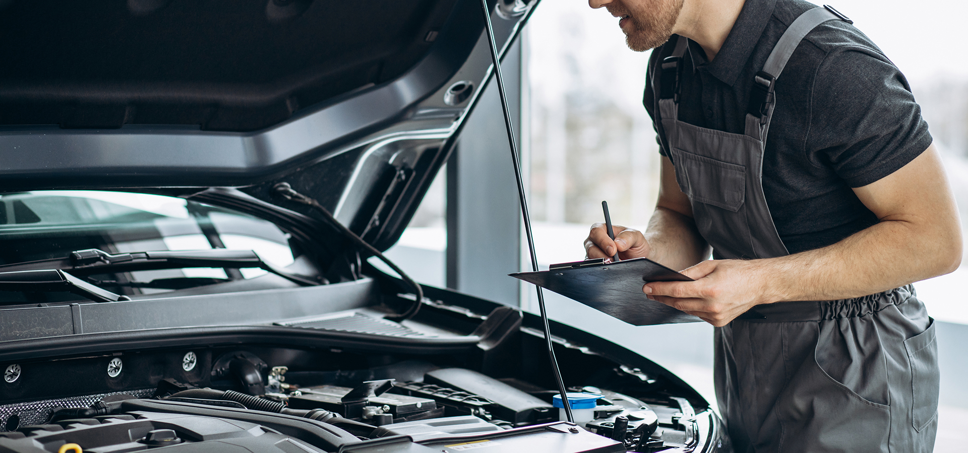 A Telle Tire mechanic holding a clipboard, checking off a list as he visually examines the components of a vehicle's engine.