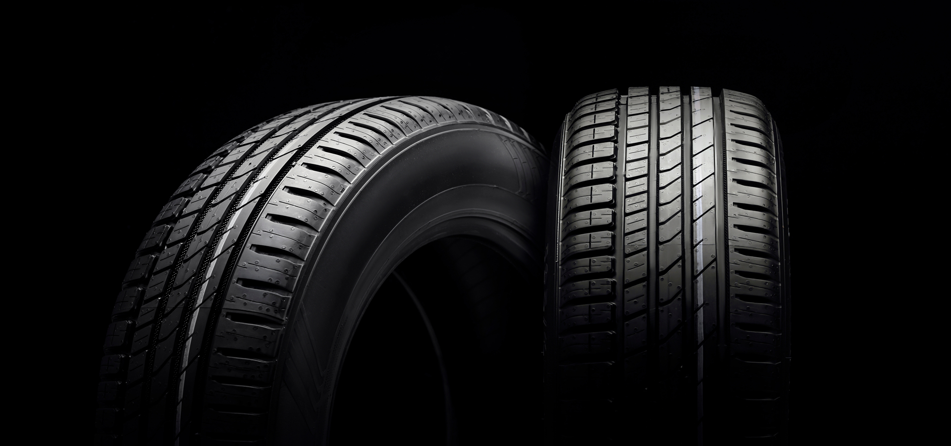 Two car tires, side-by-side on a black background lit by soft overhead lighting.